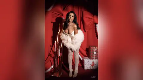 Glamorous woman in a white feathered outfit posing by lit candles in a festive red holiday setting.