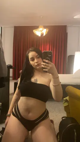 LeahBrookss poses in a black tube top and matching cut-out panties, showing her long dark hair and chest tattoo in a bedroom setting.
