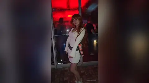 RossiNoleen stands in a white long-sleeved dress with long brown hair in a dimly lit setting.