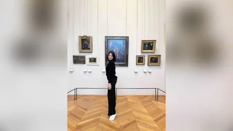 Woman in a black outfit standing in a modern art gallery with framed paintings and wooden floors.