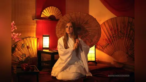Serene woman in white flowing robes sitting amidst large decorative oriental fans in warm, moody lighting.