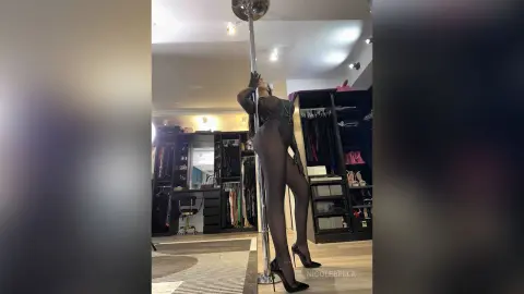 NicoleBella poses naked against a silver pole wearing a black lace bodysuit and black high heels.
