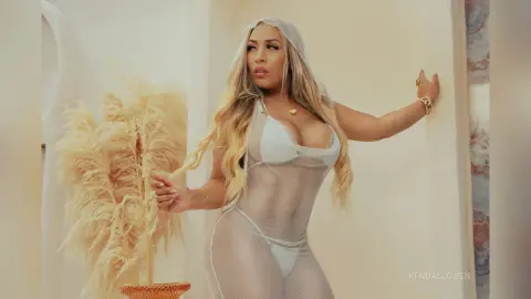 Blonde woman in a white sheer mesh bodysuit posing elegantly in a bright, minimalist indoor setting.