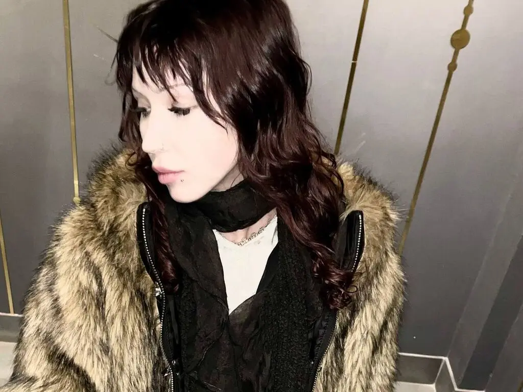 Woman with wavy dark hair wearing a cozy brown faux fur coat and black scarf in a modern indoor setting.