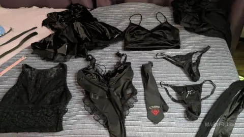 MarsyBliss displays a collection of black lace and satin lingerie, including a bodysuit, bra, skirt, and thongs, laid out on a grey bed.