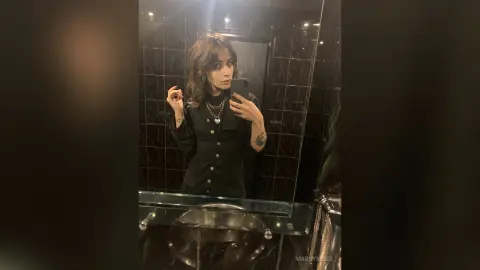 Young woman in a black long-sleeve shirt taking a moody mirror selfie in a modern dark tiled bathroom.