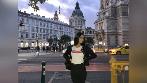 LucyMartin stands on a city street wearing a white t-shirt, black pants, and a black leather jacket with her long dark hair down.