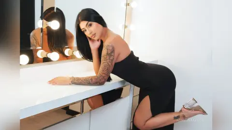 Tattooed woman in a sleek black dress posing in a bright makeup studio with a vanity mirror.