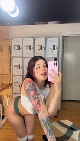 Tattooed woman in a white tank top taking a playful mirror selfie in a bright modern room.