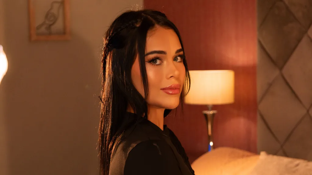 Beautiful brunette woman in a black long-sleeve top posing thoughtfully in a cozy, dimly lit bedroom.