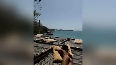 Relaxed woman sunbathing on a wooden deck overlooking a tropical turquoise ocean under a clear blue sky.