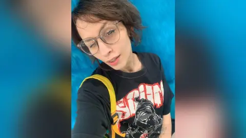 Young woman with glasses wearing a black graphic t-shirt against a bright blue background.