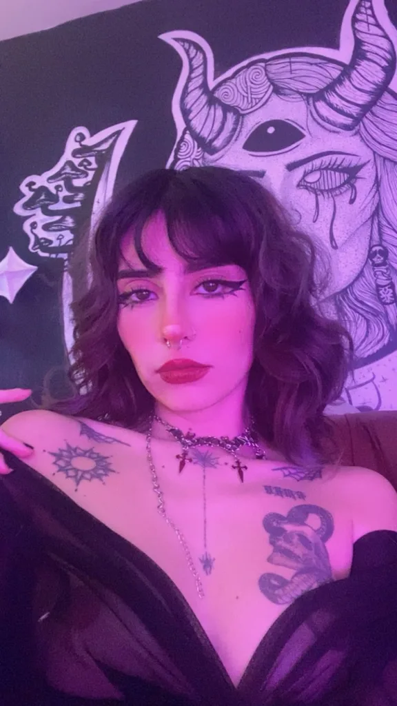 Ethlox_xxx with blue hair and heavy eyeliner poses in a spiked choker and silver necklaces, showing her chest tattoos.