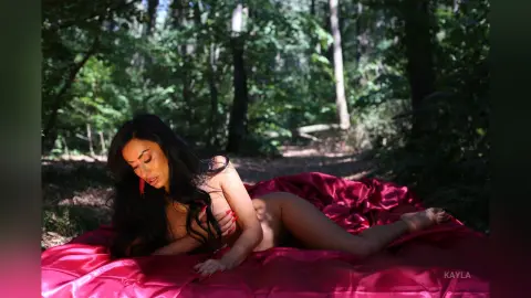Kayla lies naked on a red satin sheet in a forest, showing her long dark hair and bare body.
