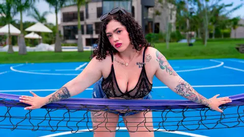 Curvy woman with tattoos in a black lace bralette relaxing on a blue hammock by a sunny outdoor tennis court.
