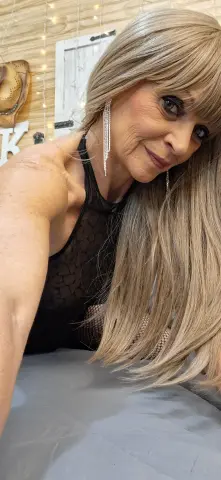 Smiling blonde woman in a black lace halter top posing in a cozy room with fairy lights.