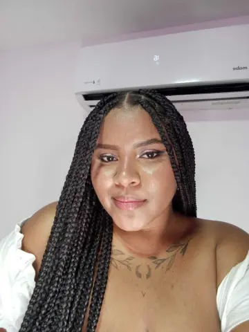 Woman with long black box braids and a white off-the-shoulder top posing in a bright indoor setting.