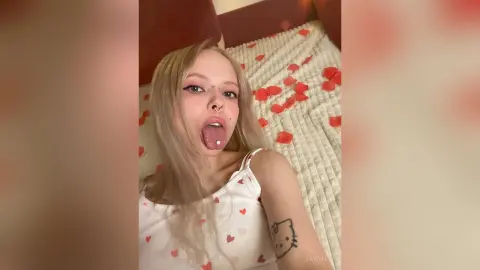 LucyLoy sticks her tongue out while lying on a bed covered in red rose petals, wearing a white tank top with red hearts.