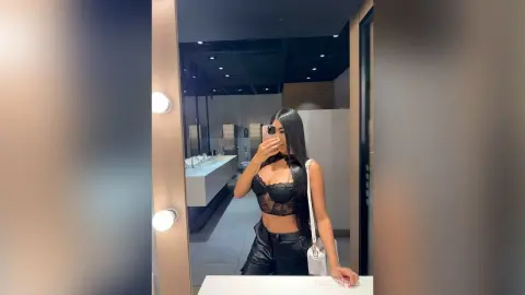 Young woman in a black lace bralette and leather pants taking a mirror selfie in a modern restroom.