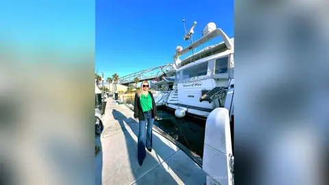 MilenaBia stands on a concrete pier wearing a green top and blue jeans, posing next to a large white yacht under a clear blue sky.