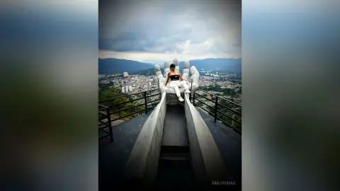 Woman in black top and white pants sitting on a giant hand sculpture overlooking a scenic mountain city view.