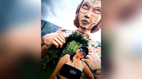 Smiling woman in sunglasses and black top posing in front of a giant earth goddess statue under a cloudy sky.