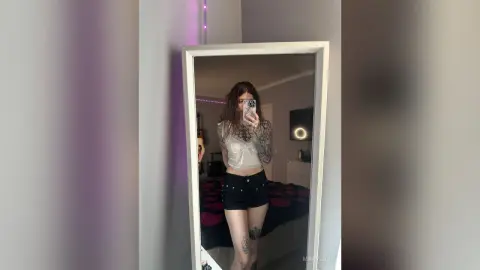 MaryLico takes a mirror selfie wearing a white crop top and black denim shorts, showing her tattoos and long dark hair.
