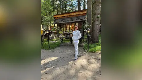 Young woman in white loungewear standing on a stone path outside a rustic wooden cabin in a sunny forest.