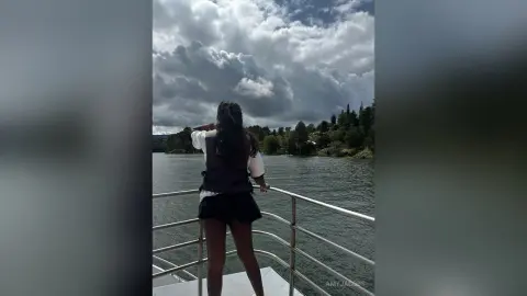 Woman in a white shirt and black shorts standing on a balcony overlooking a lake under a cloudy sky.