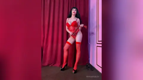 ErikaBradley poses in a red lace bodysuit and red thigh-high stockings, holding a red whip against a red curtain background.