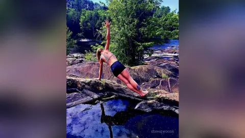 Athletic woman in black activewear performing a yoga pose on rocky riverbanks near a peaceful forest stream.