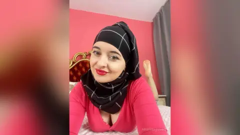 Smiling woman in a black hijab and pink top relaxing in a bright pink bedroom.