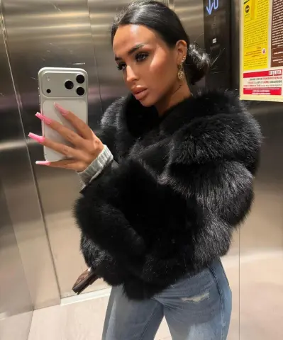 Glamorous woman in a black faux fur coat and jeans taking a mirror selfie in a modern elevator.