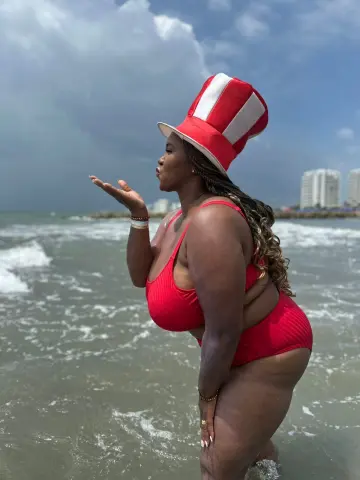 Curvy woman in a red bikini and striped top hat blowing a kiss at the beach under a cloudy sky.