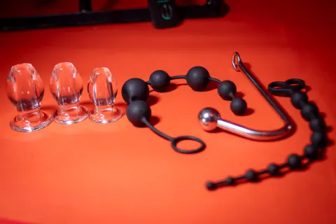 Three clear glass nipple clamps and a black beaded anal bead toy on a vibrant red background.