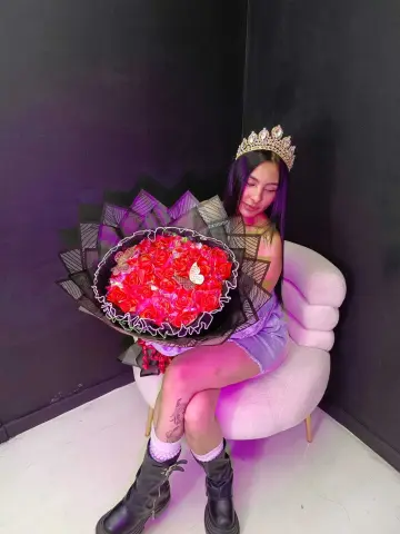 MorganHarper wears a gold crown while sitting in a white chair, holding a large bouquet of red roses against her body.