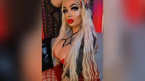 Glamorous blonde woman with tattoos in a black outfit posing with bold red lips and dramatic makeup.