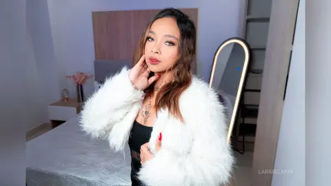 Glamorous woman in a white faux fur coat posing in a modern bedroom with soft lighting.
