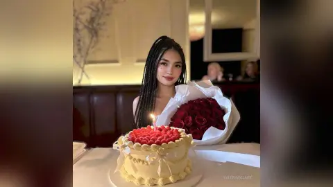ValeriaEclaire poses behind a white birthday cake with red frosting and a lit candle, holding a large bouquet of red roses.