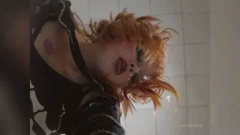 Redheaded woman with glasses in a black patterned top posing in a bright, tiled room with a moody aesthetic.