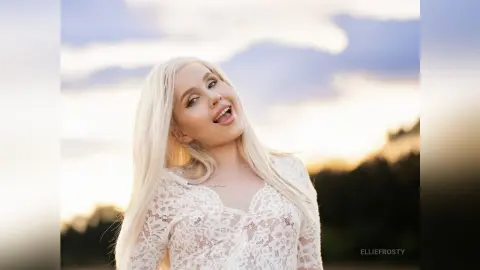 Smiling blonde woman in a white lace blouse posing outdoors during a golden hour sunset.