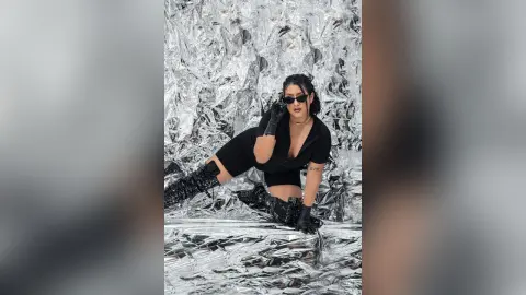 KimRoberth poses in a black outfit and sunglasses against a shiny silver foil background.