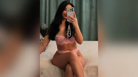 Woman in pink lace lingerie taking a mirror selfie on a bed in a cozy bedroom setting.