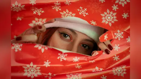 ArinnaGreen peeks through a red snowflake-patterned paper, showing her eyes and hair with red nail polish.