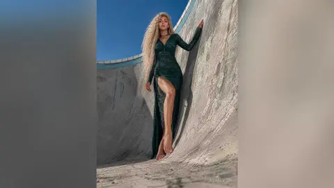 Blonde woman in a long dark green lace gown posing elegantly inside a concrete skate bowl under a blue sky.