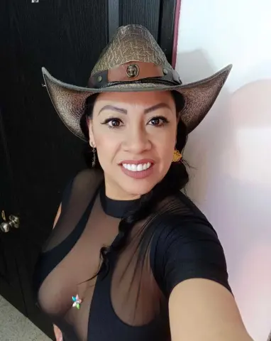 Smiling woman in a black sheer top and brown cowboy hat posing indoors with a confident mood.