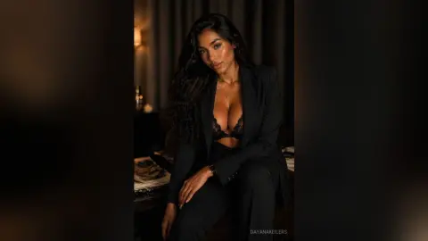 Elegant woman in a black blazer and lace lingerie posing in a moody, dimly lit indoor setting.