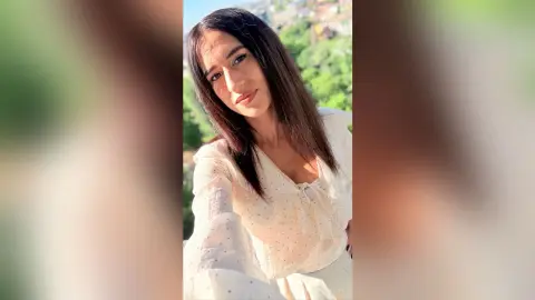 BellaLeen poses outdoors wearing a white, long-sleeved blouse with a tie-front detail and her long dark hair down.