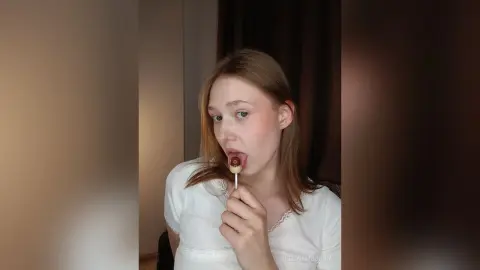 Young woman in a white blouse eating a chocolate lollipop in a cozy indoor setting.