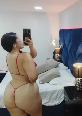 Curvy woman in red string bikini posing in a modern bedroom with blue velvet headboard.
